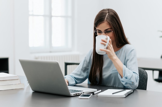 Jolie femme brune travaillant sur son ordinateur portable et buvant du café dans son bureau.