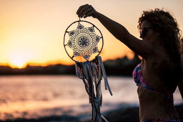 Jolie Femme Caucasienne D'âge Moyen Prendre Un Dreamcatcher Fait à La Main. Plage En Plein Air Coucher De Soleil Coloré Activité De Loisirs Pittoresque. Concept De Vacances Et D'envie De Voyager. Cheveux Bouclés Et Lunettes De Soleil Pour Voyager L