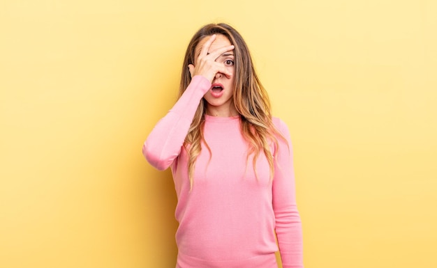 Jolie femme caucasienne ayant l'air choquée, effrayée ou terrifiée, couvrant le visage avec la main et regardant entre les doigts