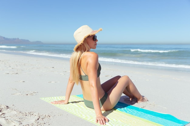 Jolie femme caucasienne blonde profitant du temps à la plage par une journée ensoleillée, portant un chapeau, assise sur une serviette et prenant un bain de soleil, avec le ciel bleu et la mer en arrière-plan. Vacances d'été à la plage tropicale.