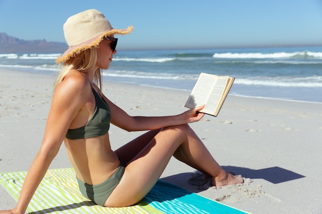 Jolie femme caucasienne blonde profitant du temps à la plage par une journée ensoleillée, portant un chapeau de soleil, assise sur une serviette et lisant, avec le ciel bleu et la mer en arrière-plan. Vacances d'été à la plage tropicale.