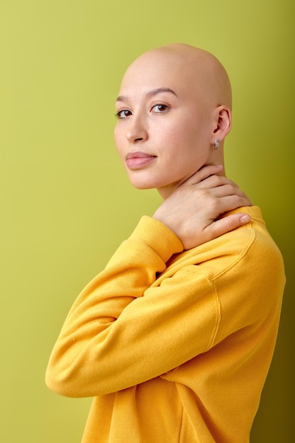 Jolie femme caucasienne chauve en chemise jaune debout isolée sur fond de studio vert, posant devant la caméra, touchant le cou. belle dame avec alopécie ou cancer. concept de soins de santé