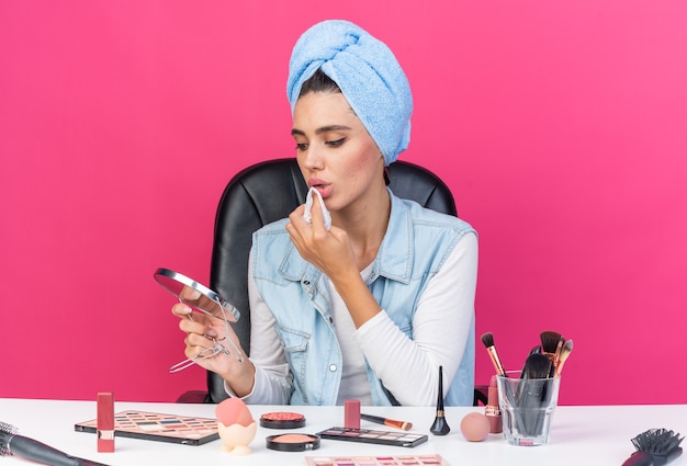 Jolie femme caucasienne confiante avec des cheveux enveloppés dans une serviette assise à table avec des outils de maquillage tenant et regardant un miroir s'essuyant la bouche avec une serviette