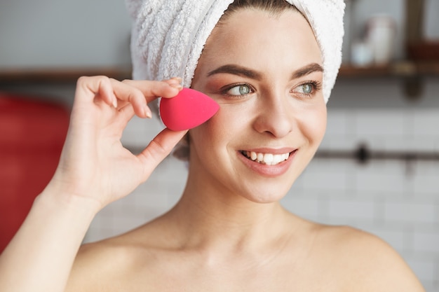 jolie femme caucasienne enveloppée dans une serviette blanche appliquant du maquillage avec une éponge cosmétique dans l'appartement