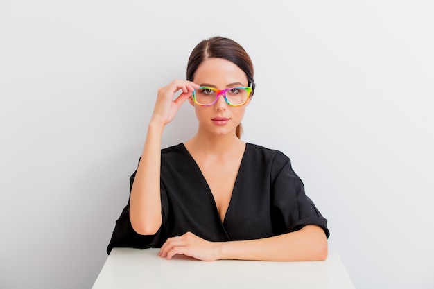 Jolie femme caucasienne rousse en robe noire et lunettes colorées assise à une table blanche dans un style lagom sur fond blanc