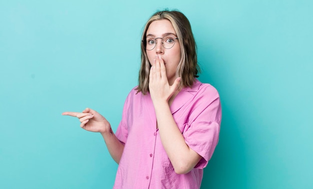 Jolie femme caucasienne se sentant heureuse choquée et surprise couvrant la bouche avec la main et pointant vers l'espace de copie latéral