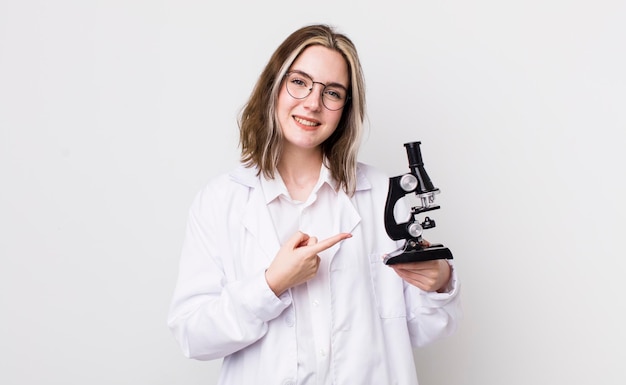 Jolie femme caucasienne souriant joyeusement se sentant heureuse et pointant vers le côté scientifique avec concept de microscope