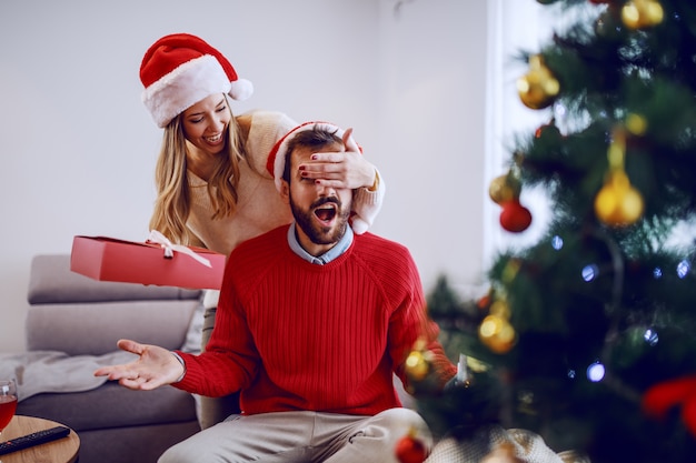 Jolie femme caucasienne souriante tenant le cadeau et couvrant les yeux de son petit ami. Les deux ayant des chapeaux de santa sur la tête. Au premier plan, l'arbre de Noël. Intérieur du salon.