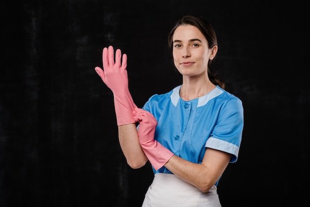 Jolie femme de chambre de mettre des gants en caoutchouc rose en se tenant debout devant la caméra sur fond noir