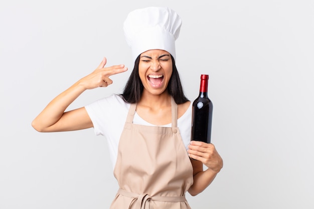 Jolie femme chef hispanique à l'air malheureuse et stressée, geste de suicide faisant un signe d'arme à feu et tenant une bouteille de vin