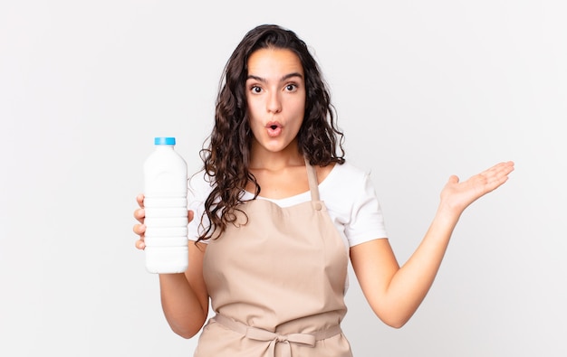 Jolie femme chef hispanique à l'air surpris et choqué, avec la mâchoire tombée tenant un objet et tenant une bouteille de lait