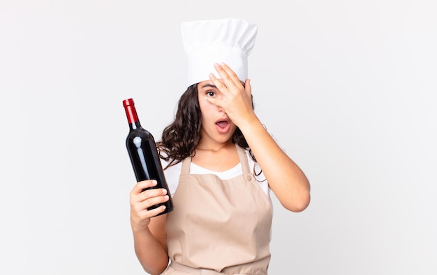 Jolie femme chef hispanique ayant l'air choquée, effrayée ou terrifiée, couvrant le visage avec la main et tenant une bouteille de vin