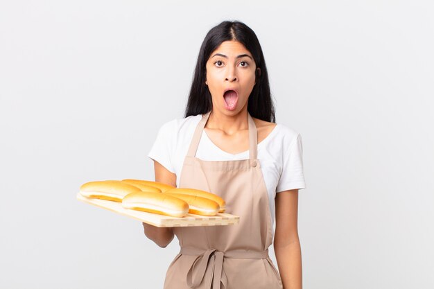 Jolie femme chef hispanique ayant l'air très choquée ou surprise et tenant un plateau avec des petits pains