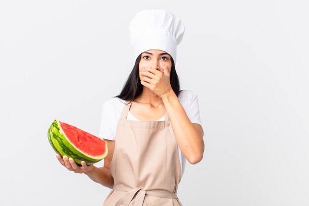 Jolie femme chef hispanique couvrant la bouche avec les mains avec un choc et tenant une pastèque