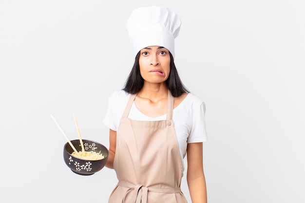 Jolie femme chef hispanique à la perplexité et à la confusion et tenant un bol de nouilles