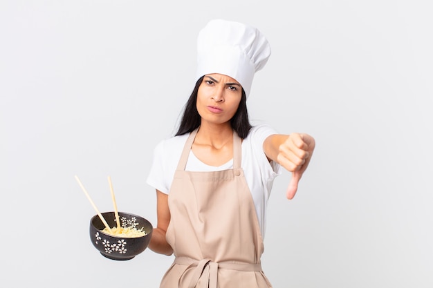 Jolie femme chef hispanique se sentant croisée, montrant les pouces vers le bas et tenant un bol de nouilles