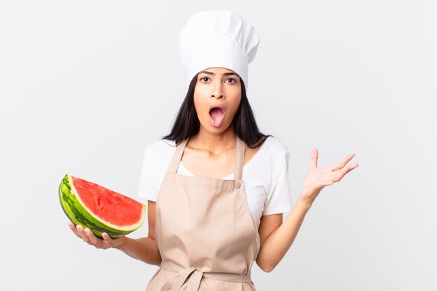 Jolie femme chef hispanique se sentant extrêmement choquée et surprise et tenant une pastèque