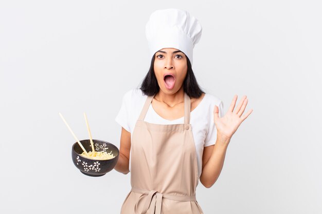 Jolie femme chef hispanique se sentant heureuse et étonnée de quelque chose d'incroyable et tenant un bol de nouilles