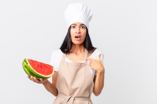 Jolie femme chef hispanique semblant choquée et surprise avec la bouche grande ouverte, pointant vers soi et tenant une pastèque