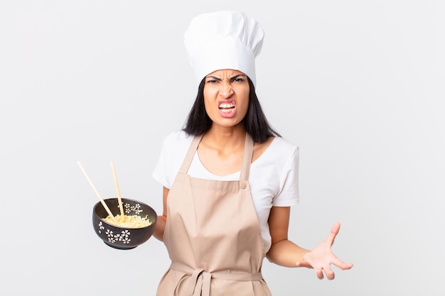 Jolie femme chef hispanique semblant en colère, agacée et frustrée et tenant un bol de nouilles