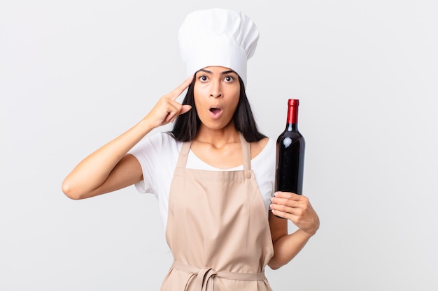 Jolie femme chef hispanique semblant surprise, réalisant une nouvelle pensée, idée ou concept et tenant une bouteille de vin