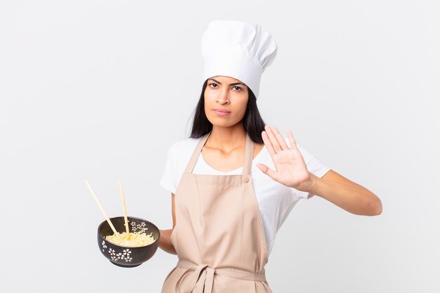 Jolie femme chef hispanique à la sérieuse montrant la paume ouverte faisant un geste d'arrêt et tenant un bol de nouilles