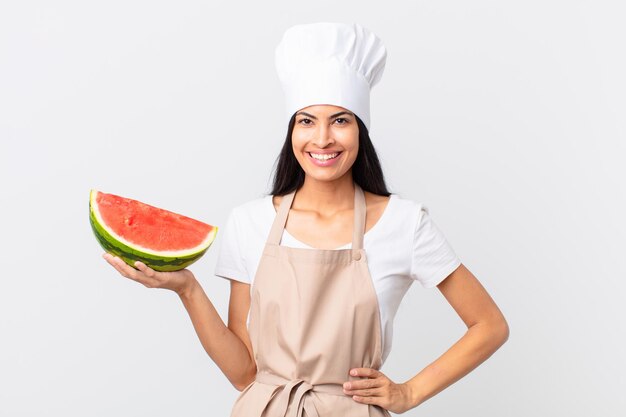 Jolie femme chef hispanique souriant joyeusement avec une main sur la hanche et confiante et tenant une pastèque