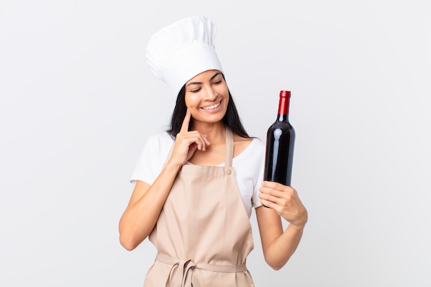 Jolie femme chef hispanique souriant joyeusement et rêvant ou doutant et tenant une bouteille de vin