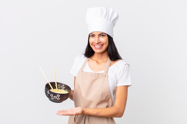 Jolie femme chef hispanique souriant joyeusement, se sentant heureuse et montrant un concept et tenant un bol de nouilles