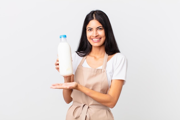 Jolie femme chef hispanique souriant joyeusement, se sentant heureuse et montrant un concept et tenant une bouteille de lait