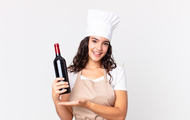 Jolie femme chef hispanique souriant joyeusement, se sentant heureuse et montrant un concept et tenant une bouteille de vin