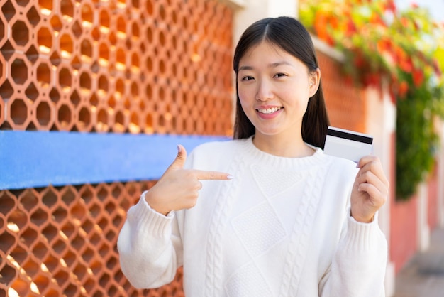 Jolie femme chinoise tenant une carte de crédit à l'extérieur avec une expression faciale surprise