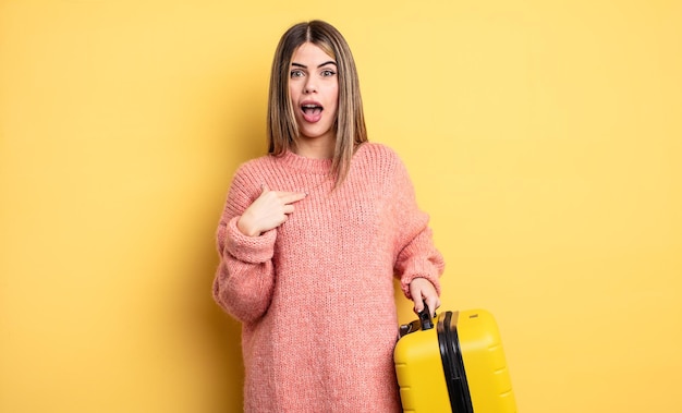 Jolie femme choquée et surprise avec la bouche grande ouverte, pointant vers soi. concept de voyage valise