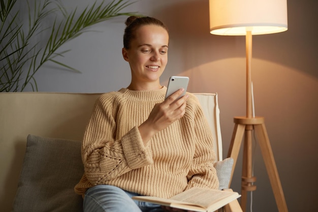 Jolie femme avec une coiffure chignon portant un pull beige assis sur un canapé dans le salon tenant un livre à la main et utilisant un téléphone portable pour passer du temps libre à lire et à vérifier les réseaux sociaux