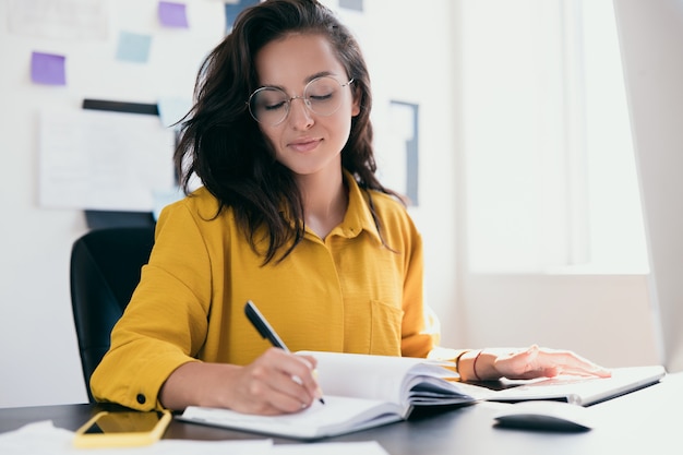 Jolie femme concentrée, implantation au bureau et plans d'écriture dans son cahier