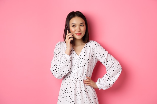 Jolie femme coréenne faisant un appel téléphonique, tenant le smartphone près de l'oreille et souriant, debout sur le rose.