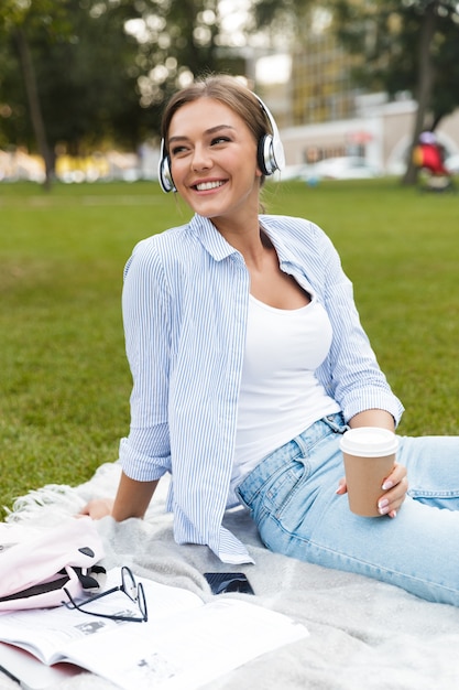 Jolie femme dans un parc à l'extérieur à l'aide d'un ordinateur portable écoutant de la musique en buvant du café.