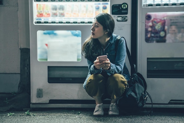 Jolie femme dans une rue sombre reposant sur un distributeur automatique de boissons moderne. jeune routard asiatique utilisant un téléphone portable attendant un ami dans une rue calme la nuit se détendre à genoux.