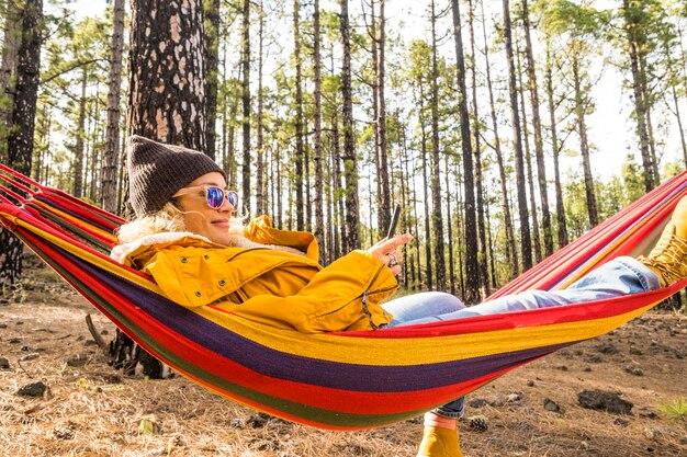 Une jolie femme détendue profite d'un hamac et d'un style de vie avec la nature autour - concept de personnes et d'environnement avec pose et repos féminins et message avec connexion téléphonique en itinérance dans la forêt