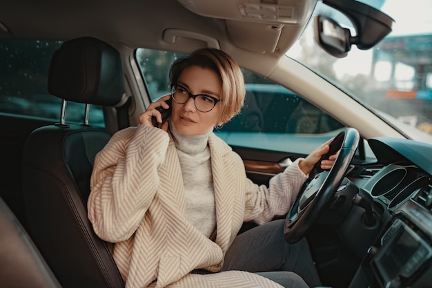 Jolie femme élégante assise dans une voiture vêtue d'un manteau d'hiver et de lunettes à l'aide d'un téléphone intelligent