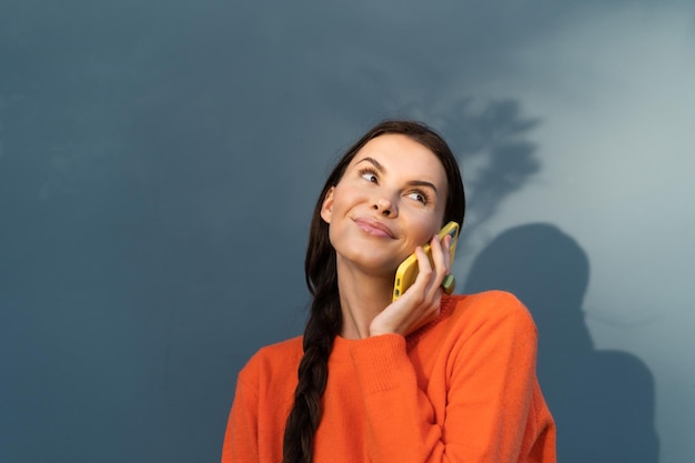 Jolie femme élégante en pull orange posant sur fond de mur bleu en plein air mignon faire un appel parlant sur téléphone mobile heureux souriant