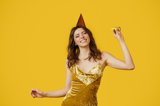 Jolie femme en élégante robe dorée et chapeau de fête profitant des vacances dansant sur studio jaune