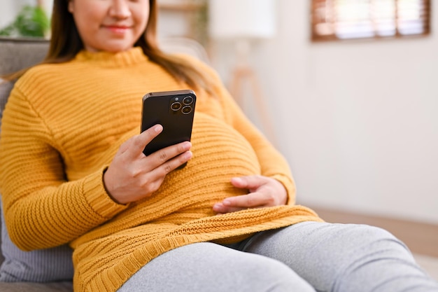 Jolie femme enceinte asiatique utilisant son smartphone sur un canapé dans le salon photo recadrée