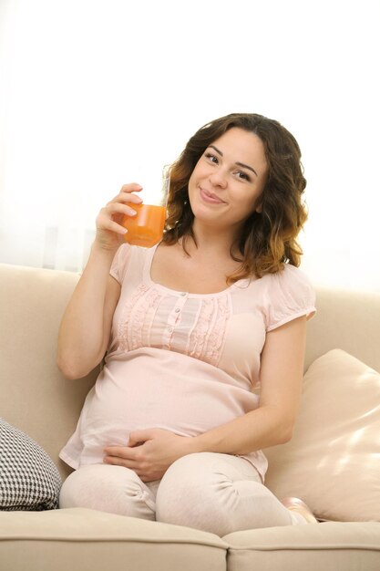 Jolie femme enceinte heureuse sur un canapé confortable avec un verre de jus dans la chambre se bouchent