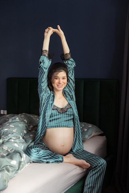 Jolie femme enceinte en pyjama est assise dans son lit et tient son ventre Derniers mois de grossesse