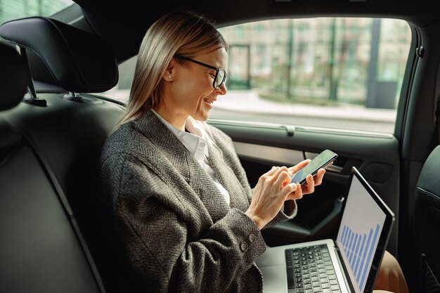 Jolie femme entrepreneur travaillant sur un ordinateur portable et utilisant le téléphone lors d'un voyage au bureau dans une voiture de luxe