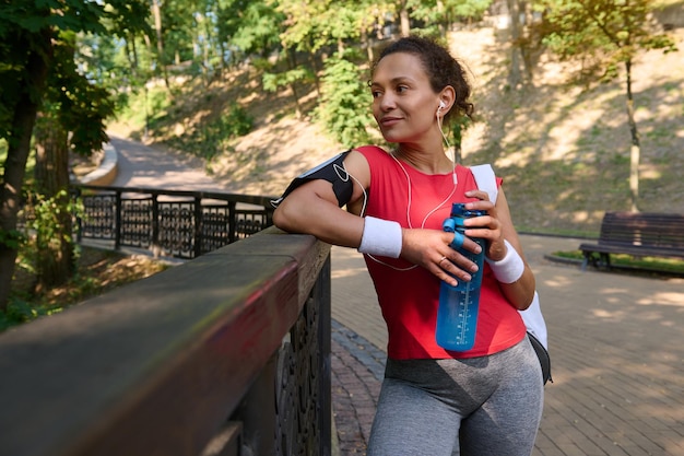 Jolie femme ethnique d'âge moyen sportive avec une bouteille d'eau relaxante dans un parc urbain après une séance d'entraînement cardio en plein air un jour d'été Sport fitness concept de mode de vie actif et sain