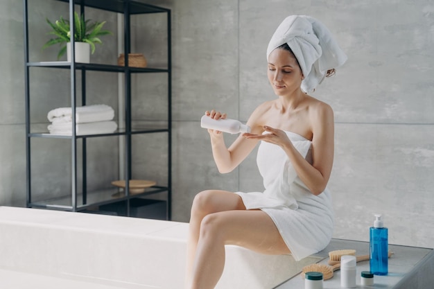 Jolie femme européenne enveloppée dans une serviette appliquant une lotion pour le corps après le bain Routine de beauté quotidienne