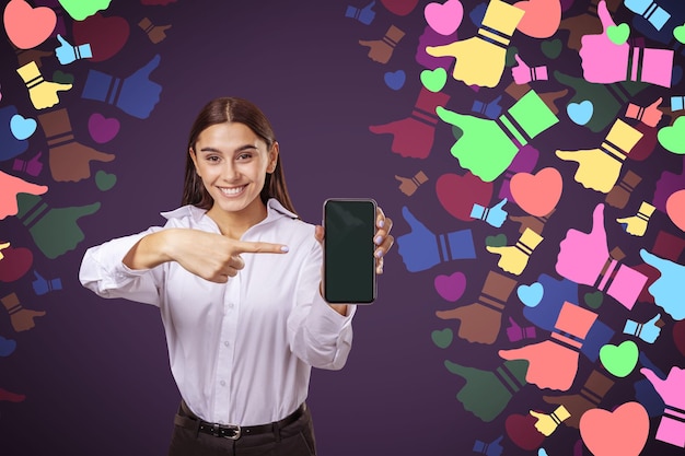 Jolie femme européenne souriante et heureuse pointant vers une maquette vide de smartphone sur des icônes abstraites colorées de médias sociaux sur fond flou Concept de rétroaction et de connexion de communication