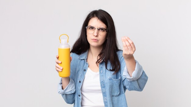 Jolie femme faisant un geste de capice ou d'argent, vous disant de payer et tenant un thermos de café